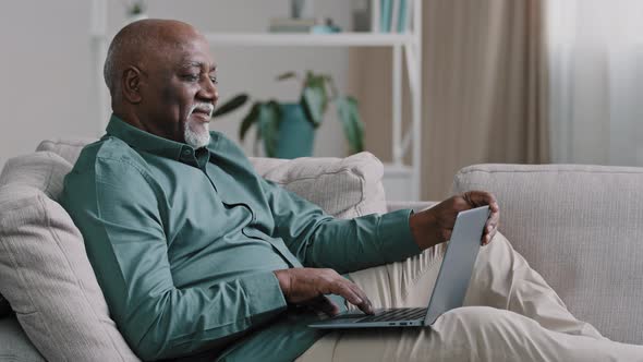 Happy Mature Pensioner Sitting on Sofa Making Internet Order Using Modern Laptop African American