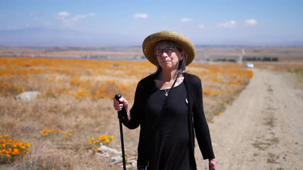 A happy middle aged woman with a hat and walking stick hiking down a dirt road with orang flowers in