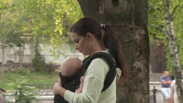 Mother with Baby Boy in Infant Kid Carrier Sling on a Walk Outdoors in City Park