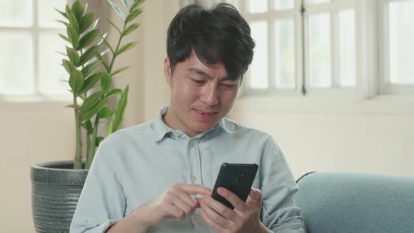 Asian Male Using Smartphone In Living Room While Lying On Sofa