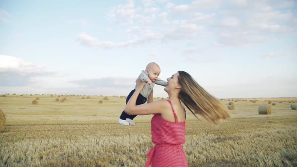 Happy Young Mother Whirling with Her Sixmonthold Son in a Field Near Haystacks