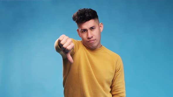 Unsatisfied man looking at camera and showing thumb down while standing on an blue background.