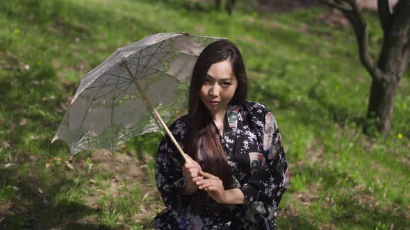 Beautiful Slim Asian Woman Looking at Camera Hiding By White Sun Umbrella Sitting on Green Spring