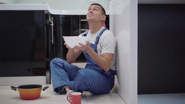 Wide Shot of Service Man in Blue Uniform Sitting on Kitchen Floor Catching Water Leaking From