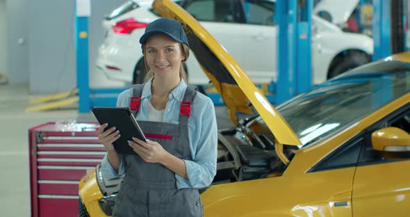 Female Car Mechanic Using Tablet Device in Big Garage with Cars. Beautiful Woman Using Tablet, Then