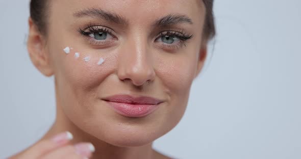 Closeup Beautiful Young Woman Applies Face Care Cream on Her Cheek Smiling for the Camera on Gray