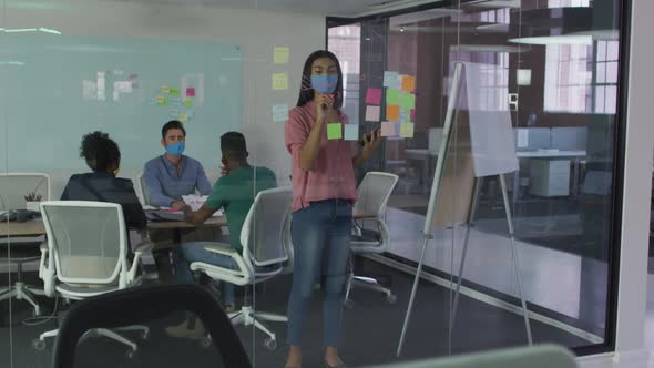 Mixed race businesswoman wearing face mask using memo notes on glass wall