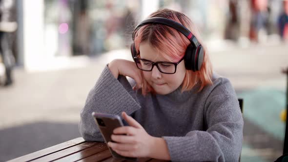 A Teenager with Pink Hair is Looking Through a Mobile Phone and Listening to Music with Headphones