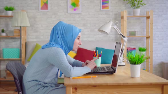 Young Muslim Woman in a Traditional Headscarf Slouches While Sitting at a Laptop and Feels Pain in