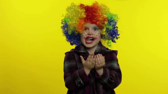 Little Child Girl Clown in Colorful Wig Hides Behind Her Hands and Shows Funny Faces. Halloween