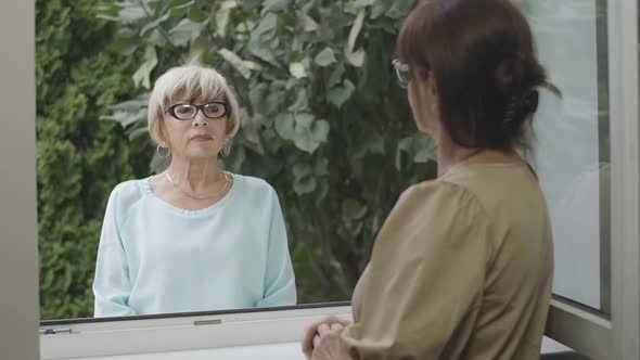 Portrait of Confident Senior Woman in Eyeglasses Standing Behind Window and Talking with Neighbor