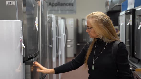 Pretty mature blonde woman looking at the features and benefits of refrigerators in a kitchen applia