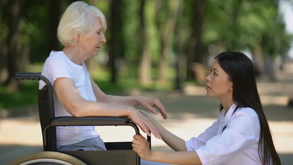 Female Volunteer Supporting Sad Elderly Woman in Wheelchair in Hospital Park