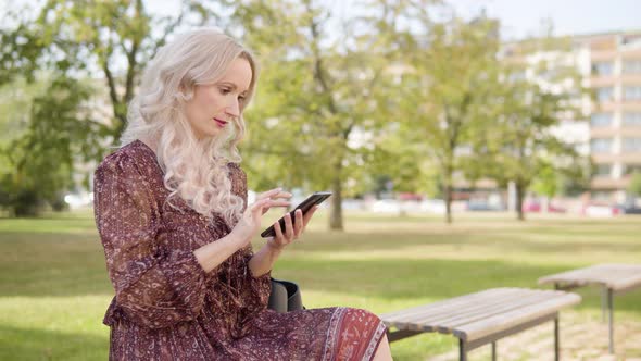 A Middleaged Caucasian Woman Works on a Smartphone As She Sits on a Bench in a Park in Urban Area