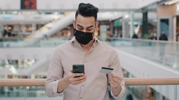 Serious Arabic Young Man in Medical Face Mask Stands in Mall Holding Credit Card Use Smartphone App