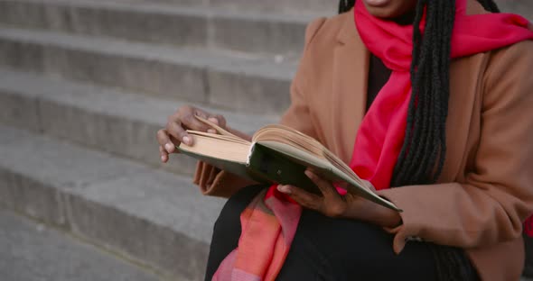 A Young Black Girl is Sitting on the Stairs on the Street and Reading a Book