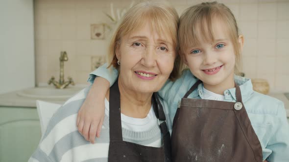 Portrait of Little Girl and Mature Woman Wearing Aprons Standing in Kitchen Hugging and Looking at