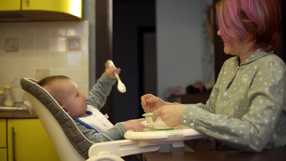 Baby is Taking Away the Spoon with Porridge While His Mother is Feeding Him and Waving It