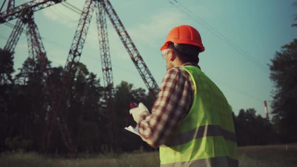 Electrical Equipment Worker Near High Voltage Tower Using Walkie Talkie and Clipboard