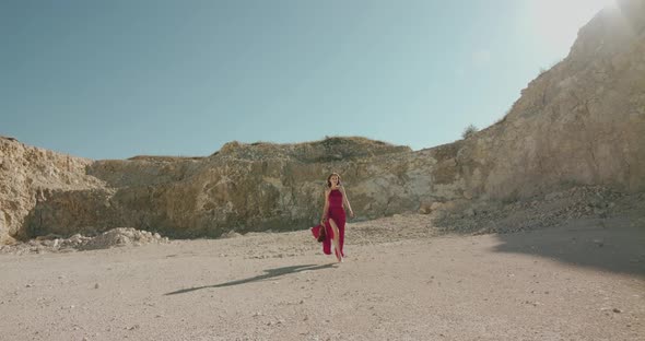 Elegant Woman in Blowing Dress Walking with Violin and Bow Among Desert at Cliff