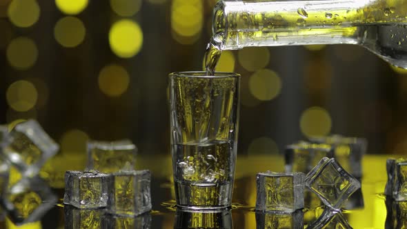 Barman Pour Frozen Vodka From Bottle Into Shot Glass. Ice Cubes Against Shiny Gold Party Background