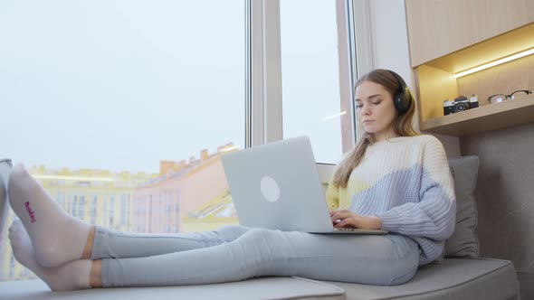 Young woman sitting by the window at home in headphones, working on a laptop. It's snowing outside.