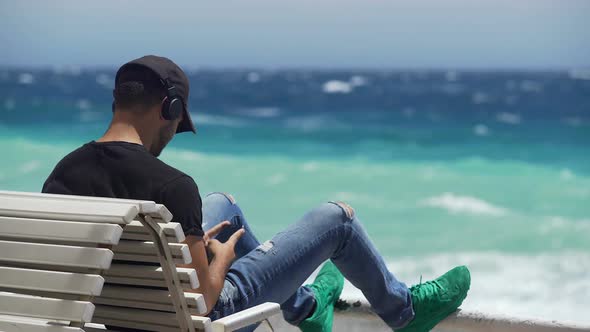 Young Guy in Headphones Sitting on Bench by Ocean Coast, Typing Message on Phone