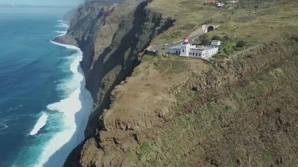 Aerial flight backwards gigantic cliff with deep ocean and lighthouse on edge during sunlight. Ponta