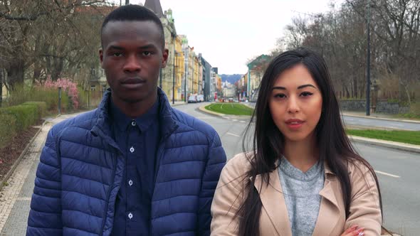 A Young Black Man and a Young Asian Woman Look at the Camera on a Sidewalk, an Urban Area