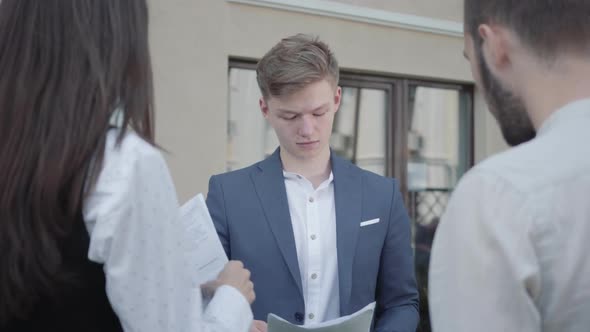 Young Woman and Two Men in Formal Wear Discussing Documents on the Terrace. The Man Giving Papers