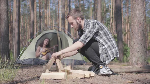 Portrait Handsome Man in a Plaid Shirt Prepares Firewood To Make a Fire Outdoors. The Girl Sits in a