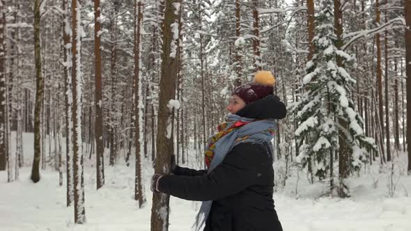 Smiling Young Woman Wearing Warm Clothes Shaking Tree and Getting Covered in Snow in Winter Forest