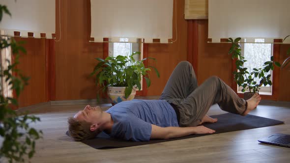 Young Adult Man Lying on Mat Doing Asana Following Instructions of Online Yoga Trainer Using Laptop