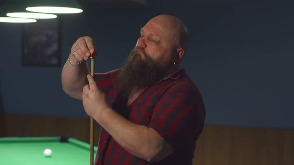 Bald brutal man with beard and a ring in his ear rubs a billiard cue with chalk, preparing for game,
