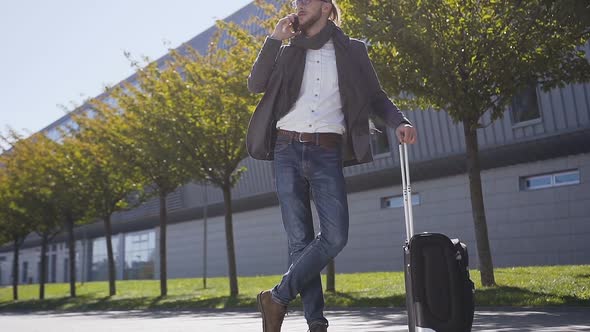 Young stylish man talking on his phone standing with suitcase 