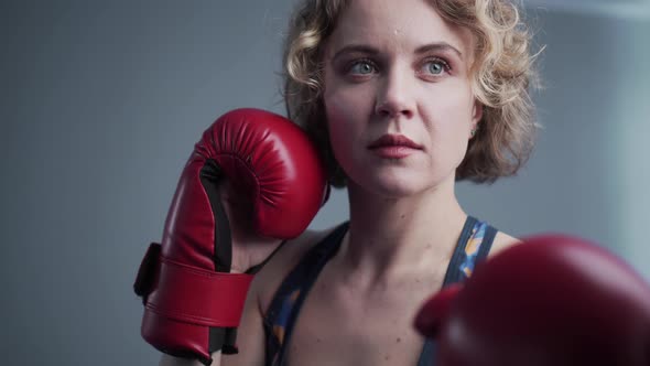 Young Beautiful Slim Woman Posing in Red Boxing Gloves in the Gym Against the Background of a Gray