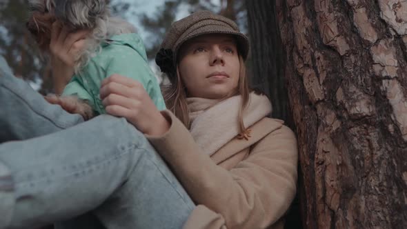 Young Woman in Coat is Sitting on Bench in Pine Forest with Dog on Her Lap Enjoying Nature