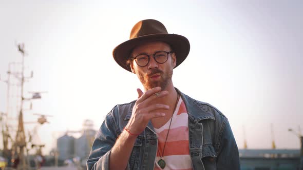 Portrait of Stylish Hipster Bearded Man in Hat and Glasses Smoking Cigarette Outdoors