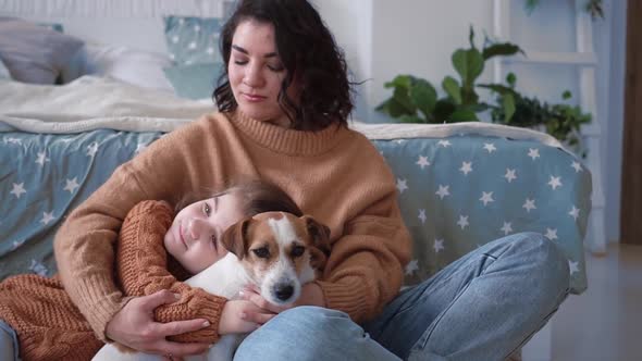 Young Mother and Daughter in Sweaters Sit on the Floor on a Rug with a Dog Jack Russell and