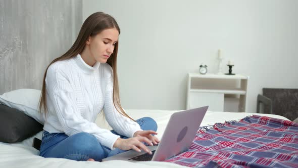 Focused Smiling Young Freelancer Woman Working Using Laptop Sitting on Bed at Cozy Home