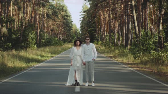 A Beautiful Hipster Couple Stands in the Middle of the Road Holding Hands and Look to the Camera