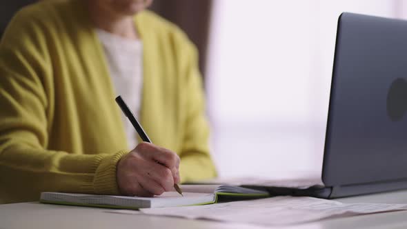 Old Lady is Learning Online with Laptop at Home Writing in Notebook Making Notes Remote Work or