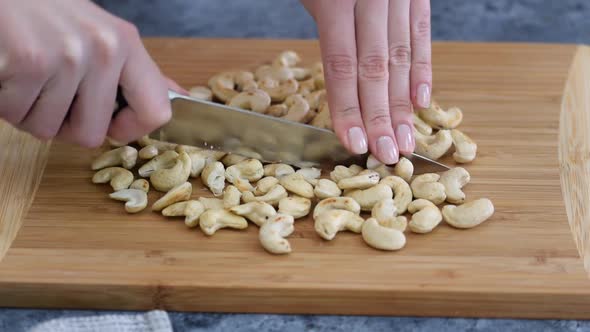 Closeup: Woman's hands with knife cut cashew nuts on a cutting board in a kitchen.