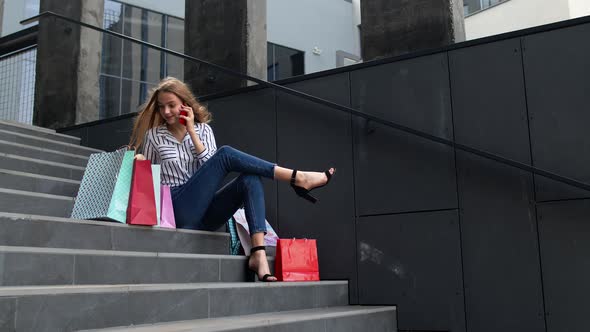 Girl Sitting on Stairs with Bags Talking on Smartphone About Sale in Shopping Mall in Black Friday
