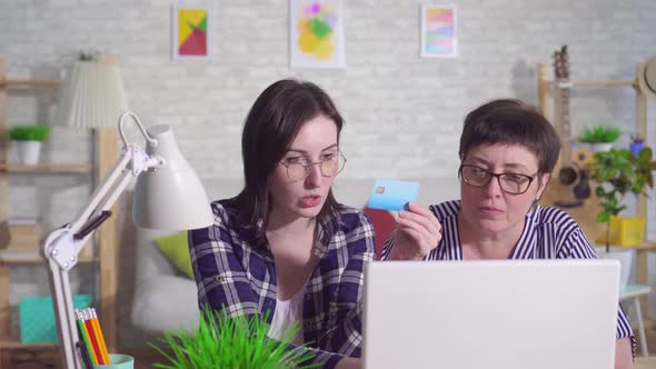 Young Woman Sitting at a Laptop Explains To a Middleaged Woman How To Use a Bank Card on the