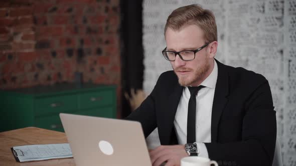 Young American Businessman Is Working on Project and Drinking Coffee at Desk in Office.