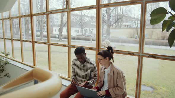 Man and Woman Discussing Project on Laptop on Window Sill