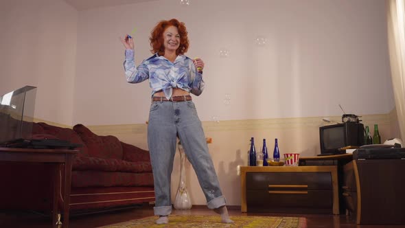 Wide Shot Joyful Adult Redhead Retro Woman Blowing Soap Bubbles Standing in Vintage Bedroom