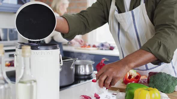 African american senior man wearing apron putting vegetable peelings in a compost bin in the kitchen