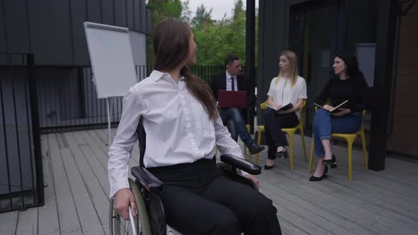 Charming Smiling Disabled Woman in Wheelchair Looking Back at Colleagues Discussing Project and